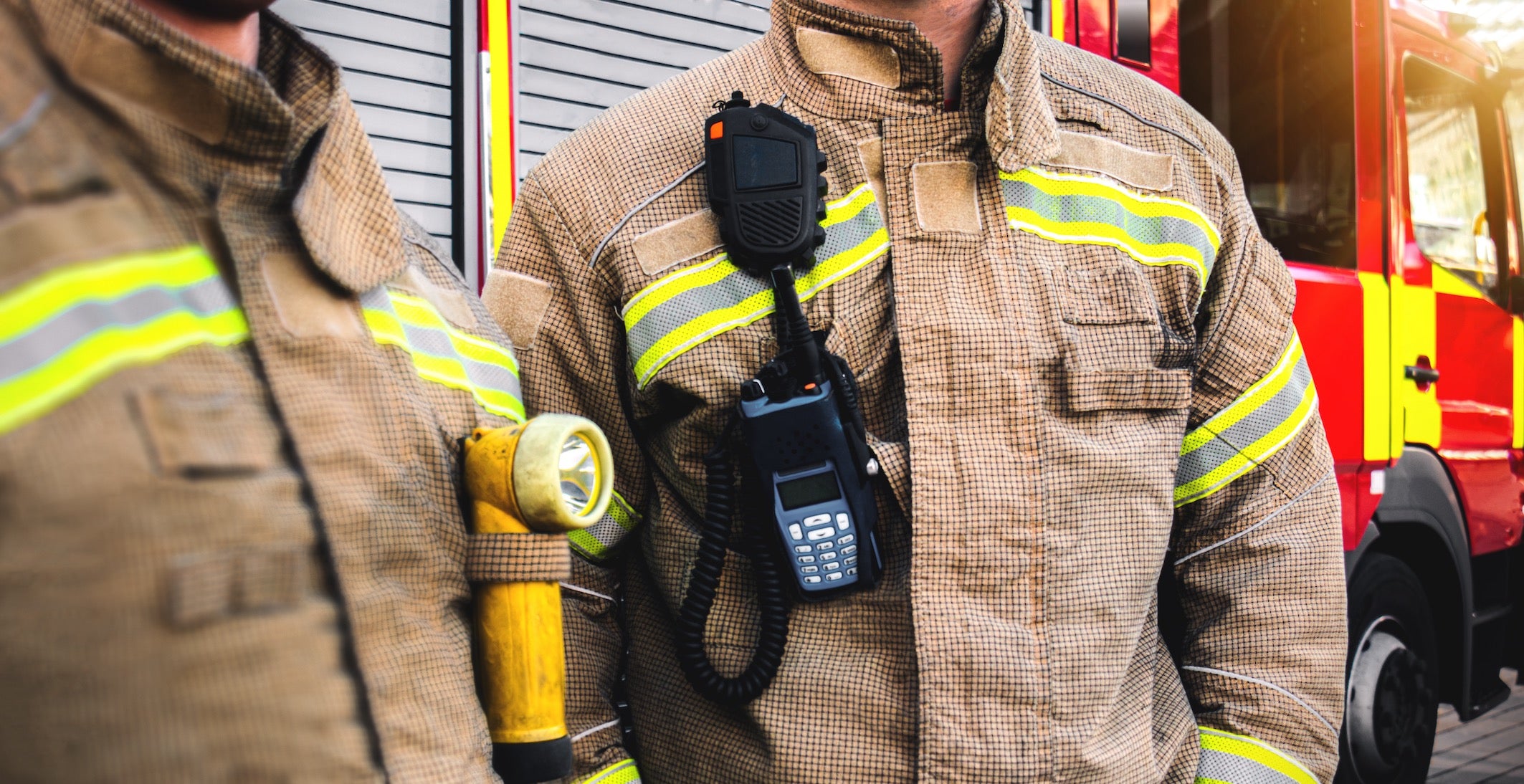 Two firefighters in protective gear with a fire engine in the background
